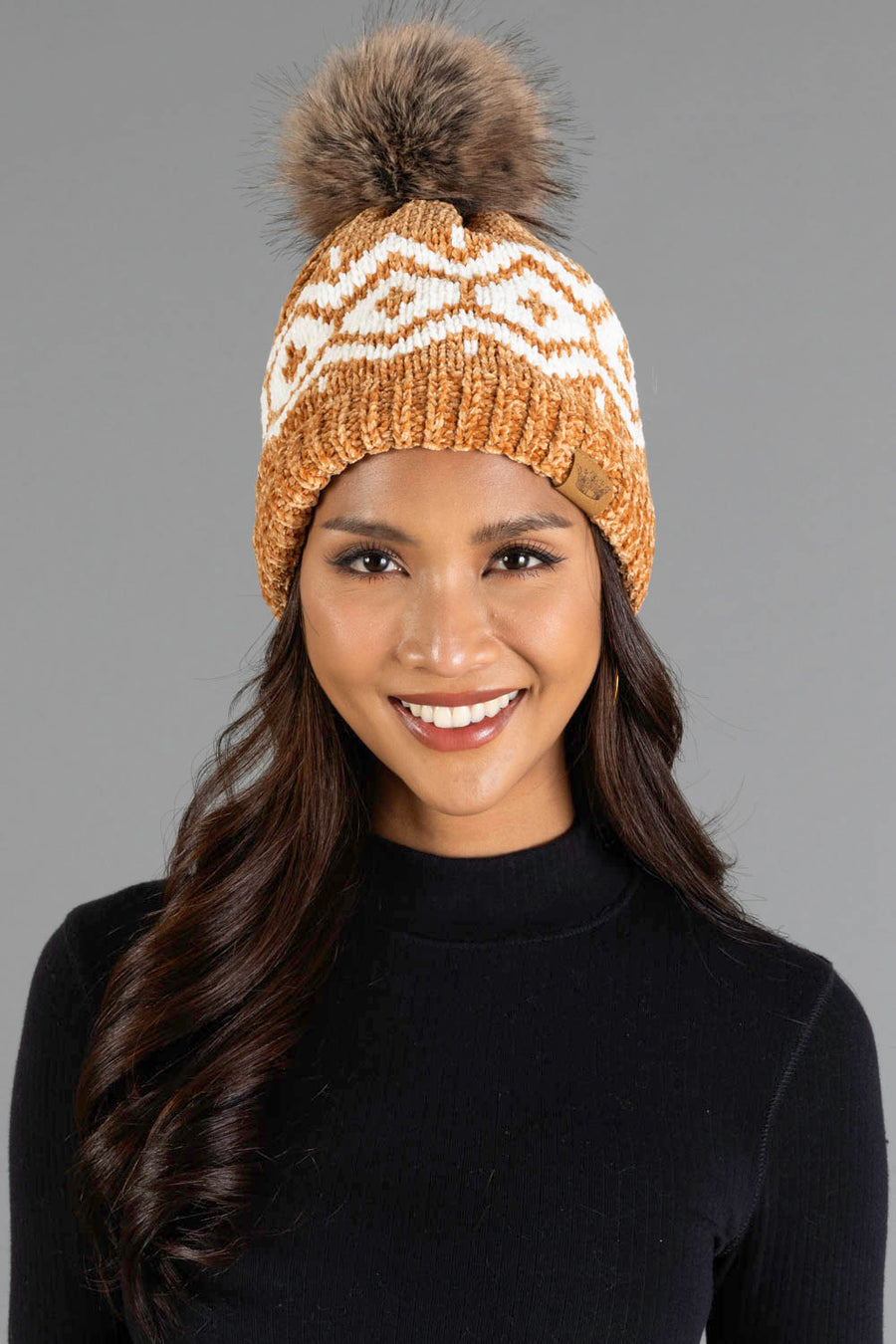 A woman smiles while wearing the Panache Apparel Collection Camel & Cream Patterned Chenille Pom Hat, featuring a cozy chenille knit with a white geometric design and a large brown pom-pom, paired with a black long-sleeve top, set against a plain gray background.