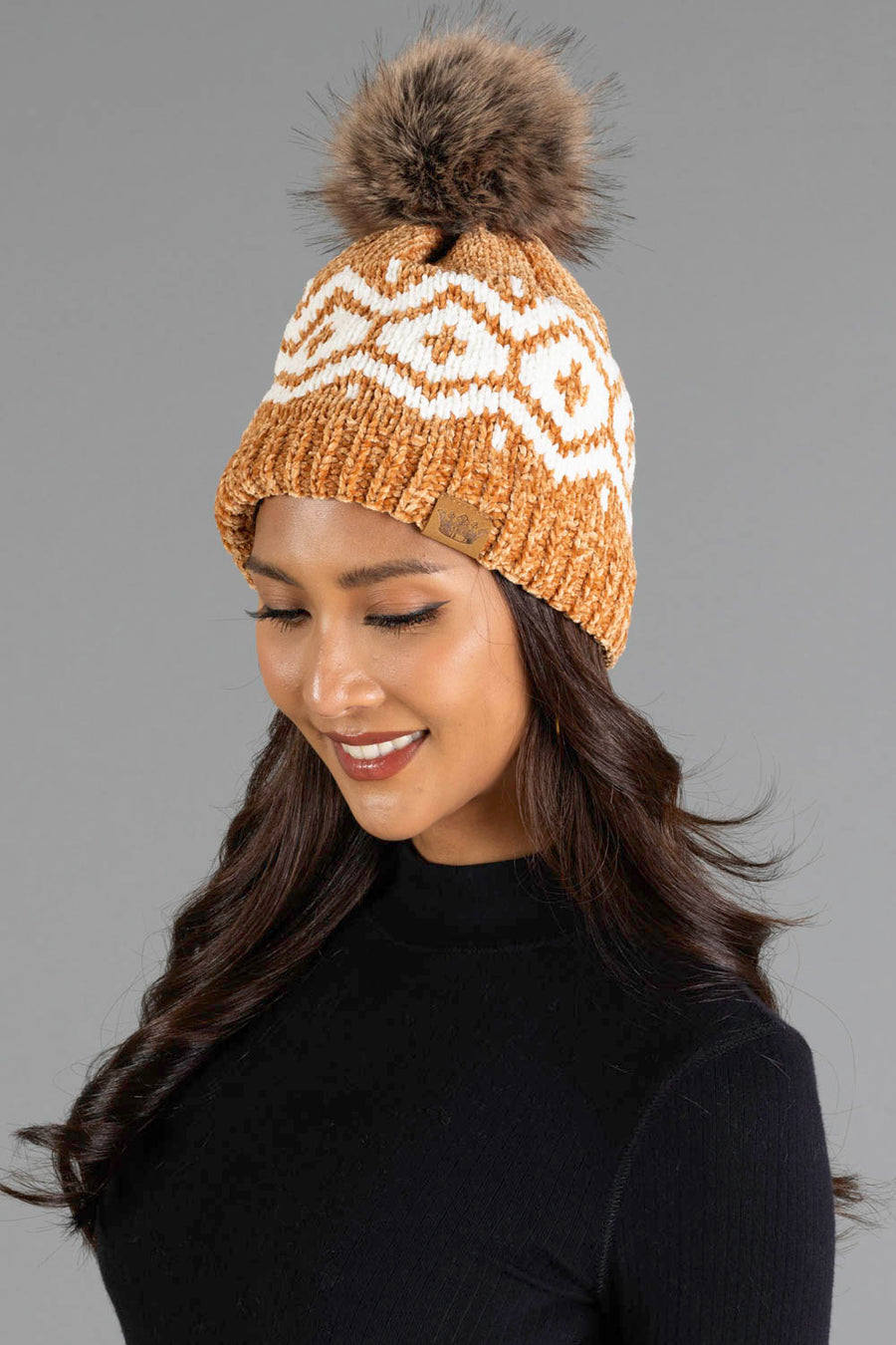 A woman with long dark hair is smiling and looking down, wearing the Panache Apparel Collection Camel & Cream Patterned Chenille Pom Hat, which features a large faux fur pom-pom, against a plain gray background.