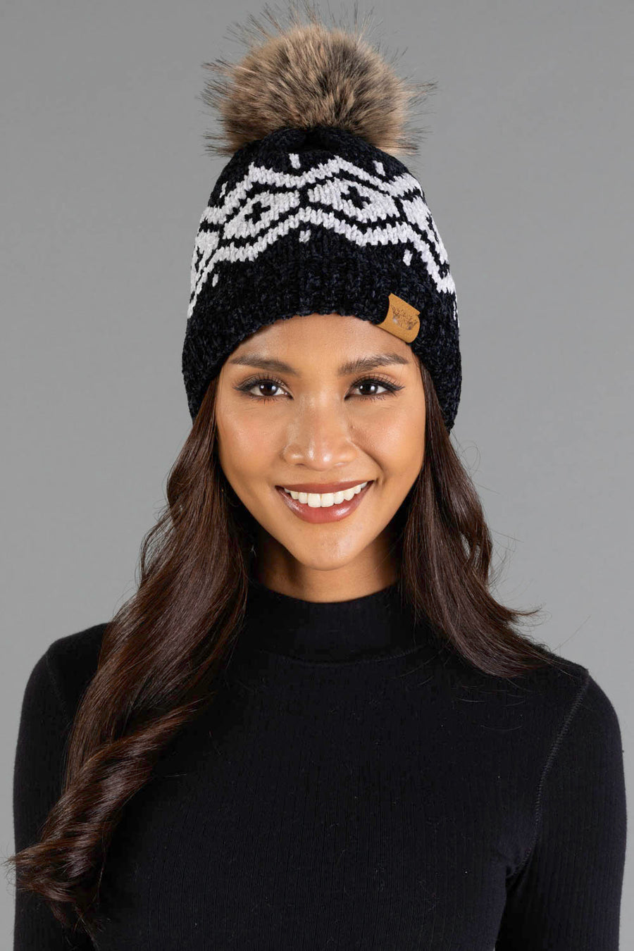 A smiling woman models the Panache Apparel Collection’s Black & Light Gray Patterned Chenille Pom Hat, featuring a white geometric design and a large faux fur pom-pom, paired with a black long-sleeve top against a plain gray background.