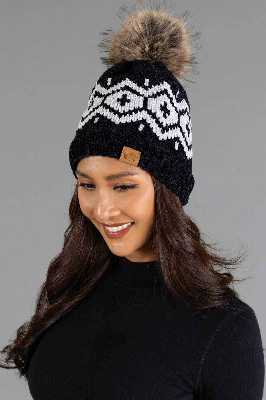 A woman with long brown hair, wearing the Panache Apparel Collection Black & Light Gray Patterned Chenille Pom Hat, featuring a large faux fur pom-pom, and a black turtleneck, smiles against a gray background.