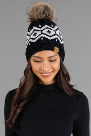 A woman with long, wavy hair smiles and looks down while wearing the Panache Apparel Collection Black & Light Gray Patterned Chenille Pom Hat, featuring a white geometric design and a brown faux fur pom-pom, against a plain gray background.