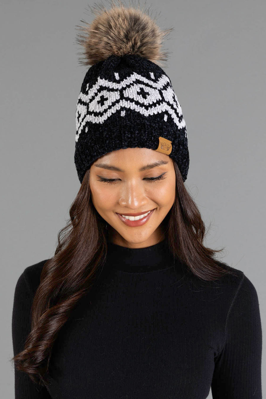 A woman with long, wavy hair smiles and looks down while wearing the Panache Apparel Collection Black & Light Gray Patterned Chenille Pom Hat, featuring a white geometric design and a brown faux fur pom-pom, against a plain gray background.