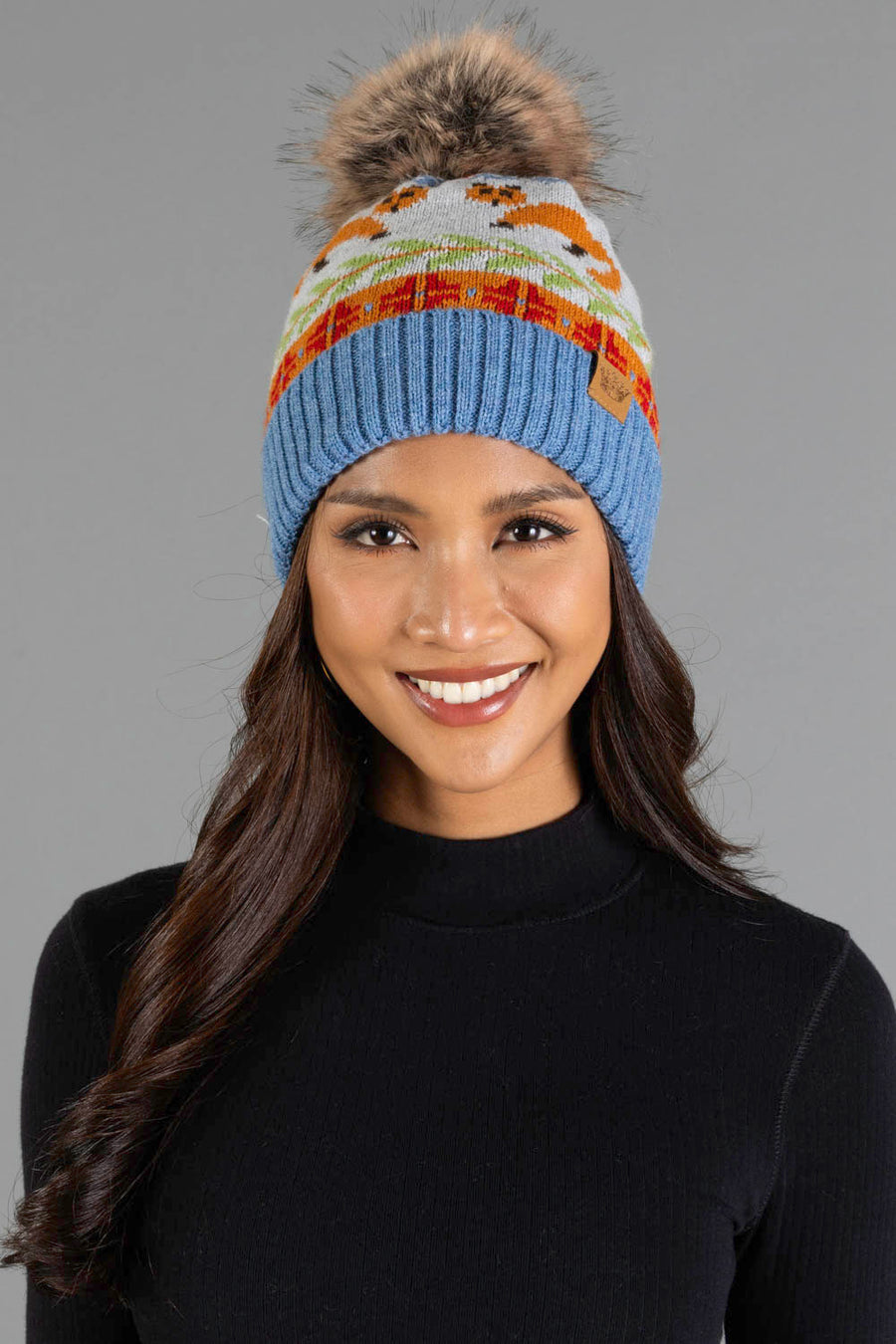 A smiling woman stands against a plain gray background, wearing Panache Apparel Collection’s Blue Fox Pattern Knit Pom Hat, a blue beanie featuring colorful patterns and a faux fur pom-pom, paired with a black sweater.