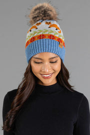 A woman smiles and looks down while wearing the Panache Apparel Collection Blue Fox Pattern Knit Pom Hat, a blue knit beanie featuring an orange fox pattern and a pom-pom, paired with a black turtleneck against a neutral gray background.