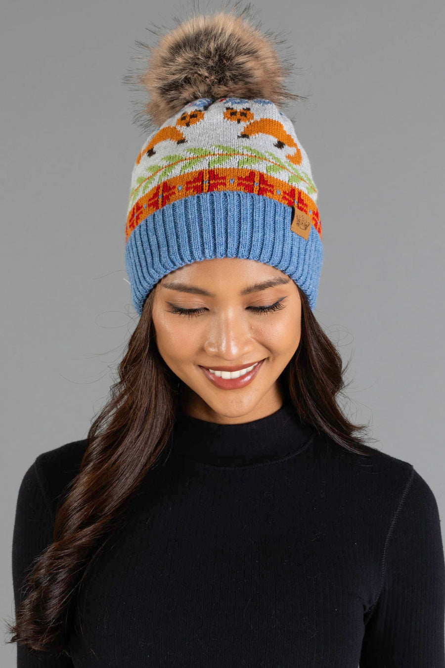A woman smiles and looks down while wearing the Panache Apparel Collection Blue Fox Pattern Knit Pom Hat, a blue knit beanie featuring an orange fox pattern and a pom-pom, paired with a black turtleneck against a neutral gray background.