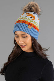 A woman smiles and looks down while wearing the Panache Apparel Collection Blue Fox Pattern Knit Pom Hat, a blue knit beanie featuring a colorful fox pattern and a faux fur pom-pom, paired with a black turtleneck against a gray background.