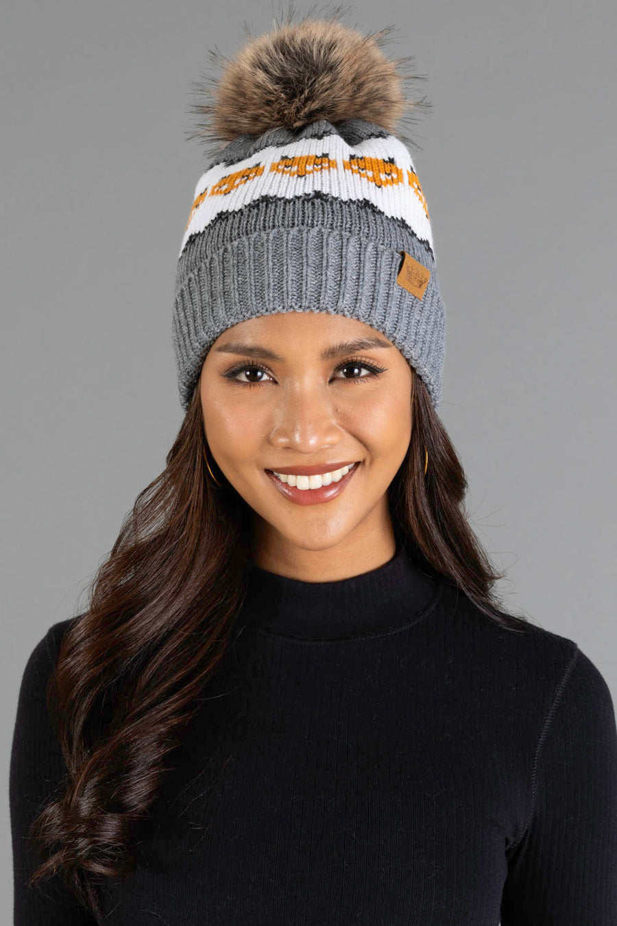 A woman smiles while wearing the Panache Apparel Collection’s Gray with Orange Fox Pattern Knit Pom Hat, styled with a black ribbed long-sleeve top against a plain gray background.