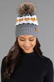 A woman with long dark hair wears the Panache Apparel Collection Gray with Orange Fox Pattern Knit Pom Hat. She is smiling and looking down, dressed in a black ribbed top against a plain gray background.