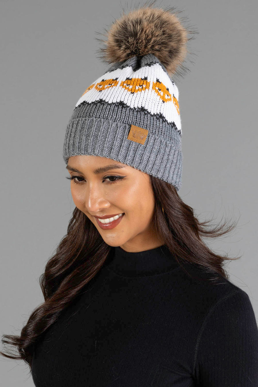A woman with long dark hair smiles while wearing the Panache Apparel Collection Gray with Orange Fox Pattern Knit Pom Hat, which features a gray knit design, playful pom-pom, white and orange fox motifs, and a leather tag. She pairs it with a black top against a gray background.