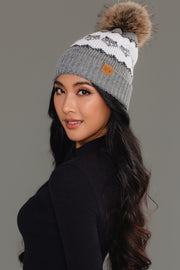 A woman with long wavy hair poses against a gray background wearing a black long-sleeve top and the Panache Apparel Collection’s Gray with Gray Fox Pattern Knit Pom Hat, a gray beanie featuring a fox pattern, pom-pom accent, and a tan brand patch.