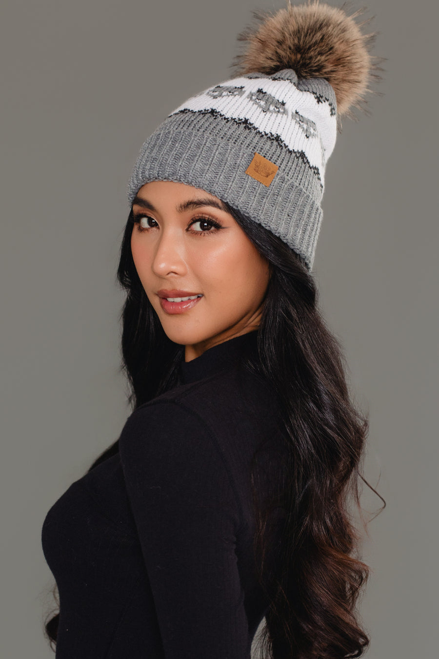 A woman with long wavy hair poses against a gray background wearing a black long-sleeve top and the Panache Apparel Collection’s Gray with Gray Fox Pattern Knit Pom Hat, a gray beanie featuring a fox pattern, pom-pom accent, and a tan brand patch.