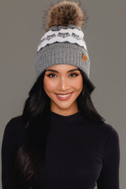 A woman with long dark hair, smiling against a plain gray background, wears the Panache Apparel Collection’s Gray with Gray Fox Pattern Knit Pom Hat, a gray beanie featuring a white fox design and brown pom-pom, paired with a black long-sleeve top.