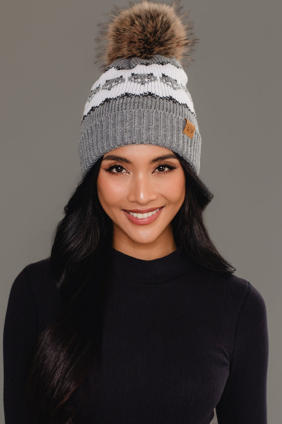 A woman with long dark hair, smiling against a plain gray background, wears the Panache Apparel Collection’s Gray with Gray Fox Pattern Knit Pom Hat, a gray beanie featuring a white fox design and brown pom-pom, paired with a black long-sleeve top.