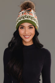 A woman with long dark hair, smiling in front of a plain gray background, wears the Panache Apparel Collection Olive & Cream Bird Pattern Knit Pom Hat and a black long-sleeve top.