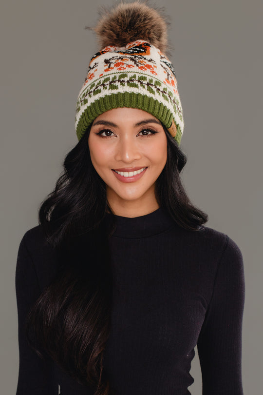 A woman with long dark hair, smiling in front of a plain gray background, wears the Panache Apparel Collection Olive & Cream Bird Pattern Knit Pom Hat and a black long-sleeve top.