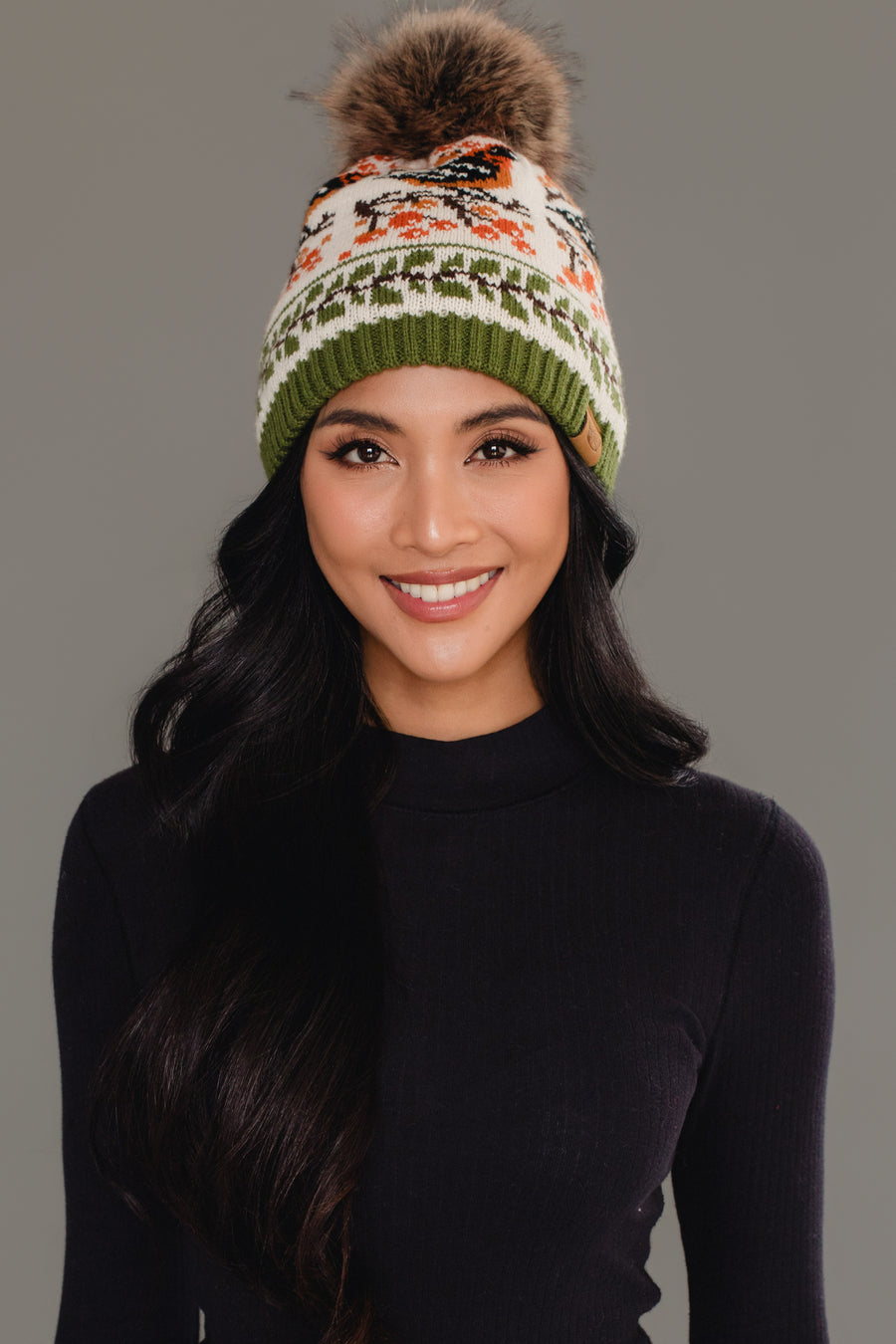 A woman with long dark hair, smiling in front of a plain gray background, wears the Panache Apparel Collection Olive & Cream Bird Pattern Knit Pom Hat and a black long-sleeve top.