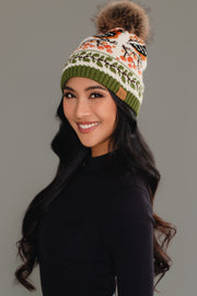 A woman with long black hair smiles against a plain gray background, wearing a black top and the Olive & Cream Bird Pattern Knit Pom Hat by Panache Apparel Collection.