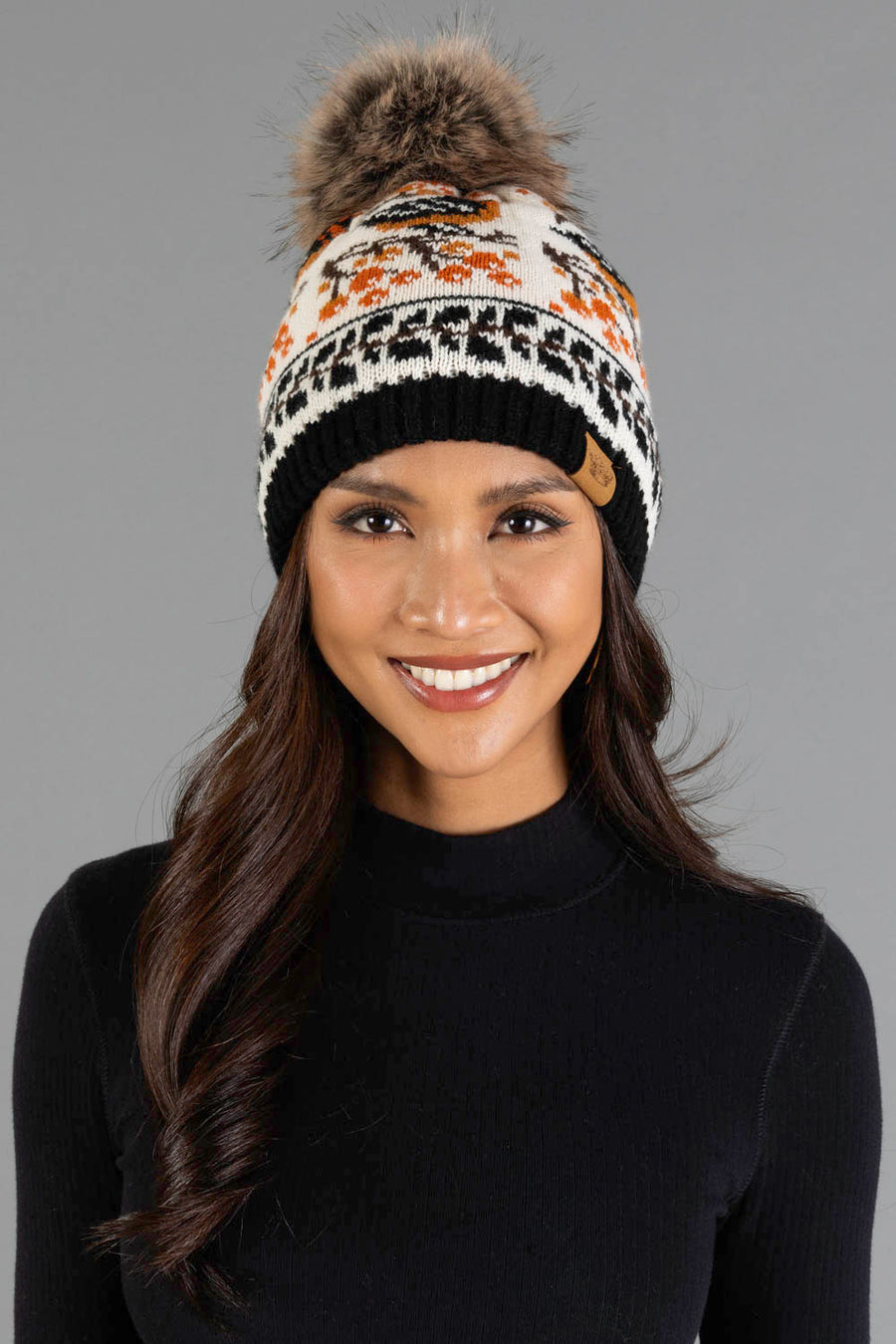 A woman smiles at the camera against a plain gray background, wearing a black turtleneck and the Panache Apparel Collection’s Black & Cream Bird Pattern Knit Pom Hat.