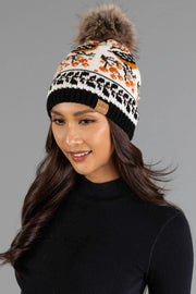 A woman with long brown hair, wearing a black turtleneck and the Panache Apparel Collection Black & Cream Bird Pattern Knit Pom Hat featuring a white, black, and orange design with a brown faux fur pom-pom, smiles against a gray background.
