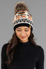 A woman with long dark hair, wearing a black turtleneck and the Panache Apparel Collection Black & Cream Bird Pattern Knit Pom Hat, featuring orange and black designs and a large faux fur pompom, smiles with her eyes closed against a gray background.