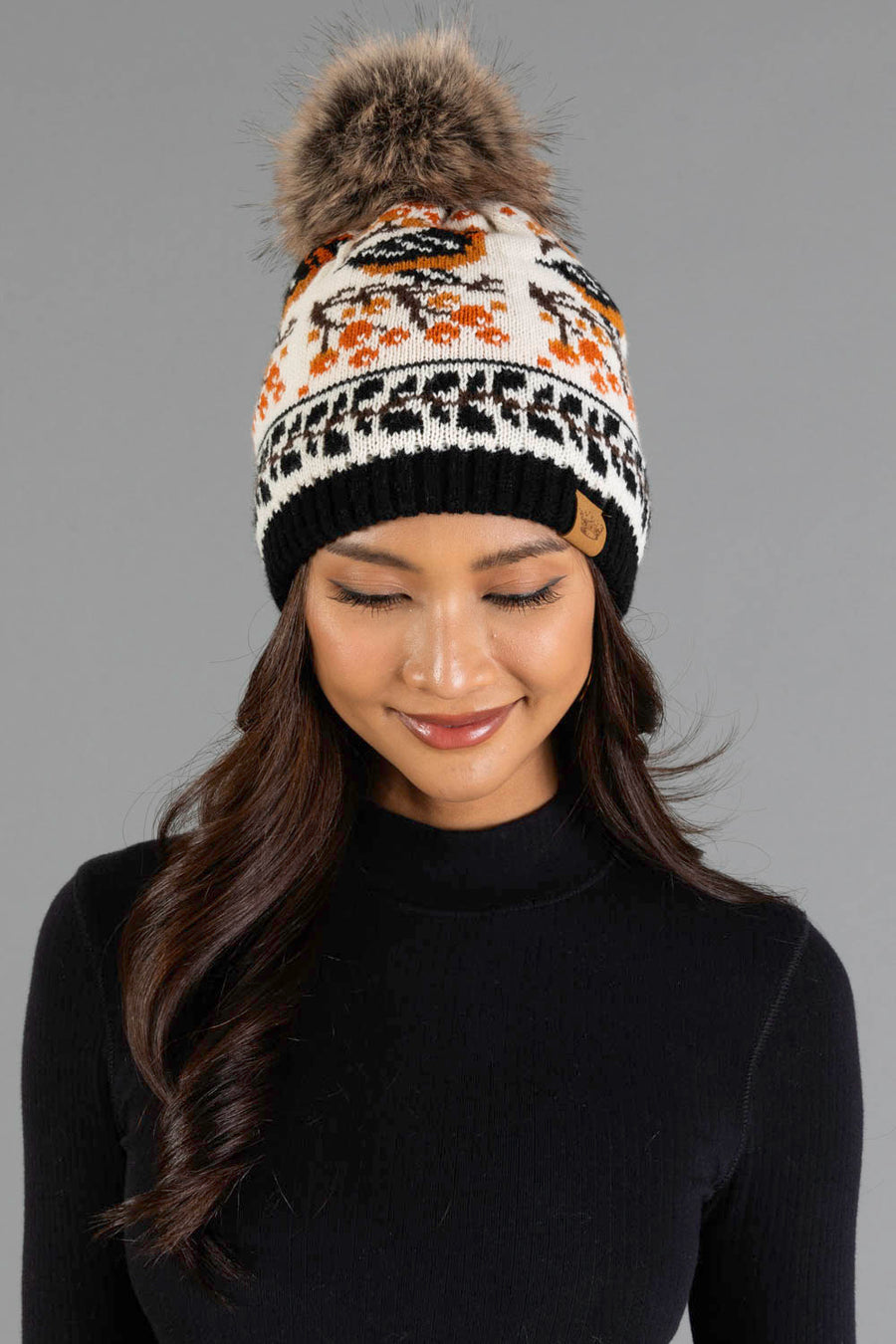 A woman with long dark hair, wearing a black turtleneck and the Panache Apparel Collection Black & Cream Bird Pattern Knit Pom Hat, featuring orange and black designs and a large faux fur pompom, smiles with her eyes closed against a gray background.