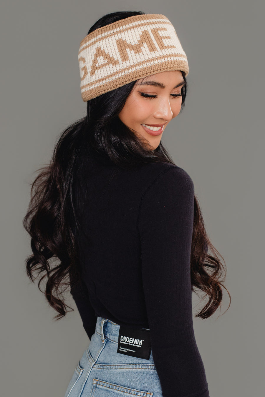 A woman with long dark hair smiles over her shoulder against a plain gray background, wearing Panache Apparel Collection’s Beige & Tan Game Day Knit Headband, a black long-sleeve shirt, and light blue jeans.