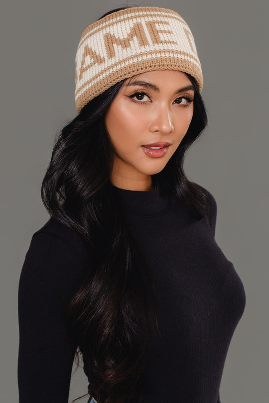 A woman with long black hair models the Panache Apparel Collection's Beige & Tan Game Day Knit Headband, featuring partial text, paired with a fitted black long-sleeve top, against a plain gray background.