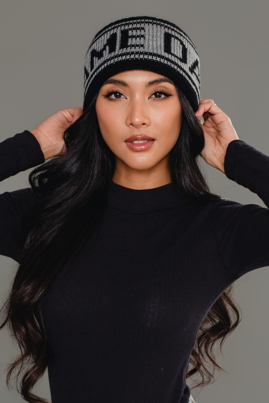 A woman with long black hair wears the Panache Apparel Collection Black & Gray Game Day Knit Headband and a black long-sleeve top, posing with her hands touching the sides of the headband against a plain gray background.