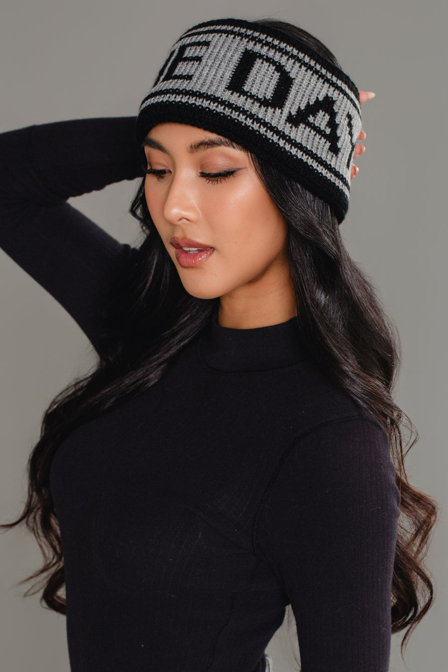 A woman with long, wavy dark hair models the Panache Apparel Collection Black & Gray Game Day Knit Headband and a fitted black top, posing with one hand behind her head against a plain gray background.