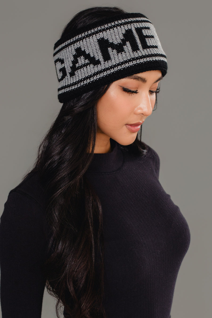 A woman with long dark hair wears a black turtleneck and the Black & Gray Game Day Knit Headband by Panache Apparel Collection, which features "GAME" across the front, as she looks down against a plain gray background.