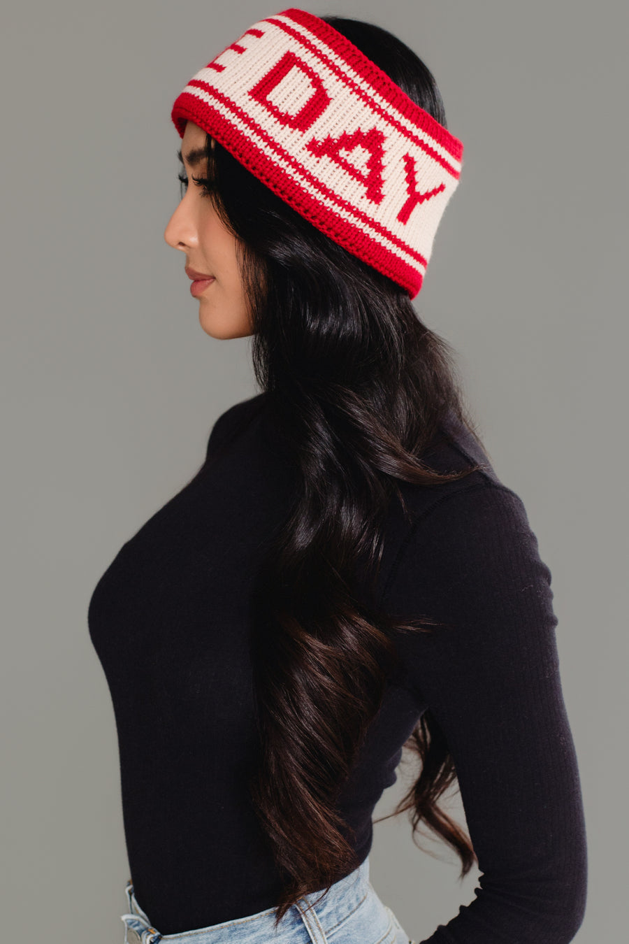A woman in profile wears the Panache Apparel Collection Red & Beige Game Day Knit Headband, styled with long wavy dark hair, a black long-sleeve top, and light blue jeans against a plain gray background.
