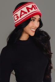 A smiling woman with long dark hair, wearing a black top and the Panache Apparel Collection Red & Beige Game Day Knit Headband—featuring "GAME" lettering—poses against a gray background.
