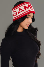 A woman with long black hair, wearing a black turtleneck and the Panache Apparel Collection Red & Beige Game Day Knit Headband featuring the word "GAME," looks down and to the side against a gray background.