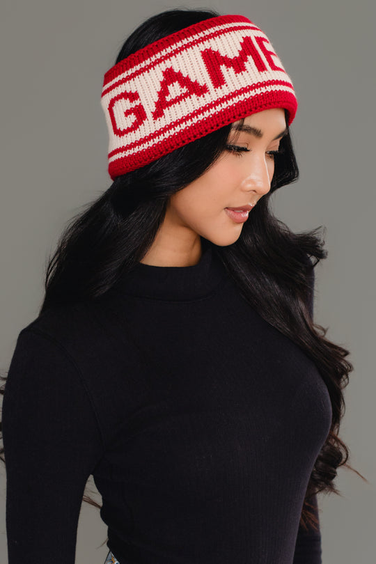 A woman with long black hair, wearing a black turtleneck and the Panache Apparel Collection Red & Beige Game Day Knit Headband featuring the word "GAME," looks down and to the side against a gray background.