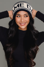 A woman with long, wavy black hair smiles while holding her Dark Navy & Beige Game Day Knit Headband from Panache Apparel Collection. She wears a black top and stands against a gray background.