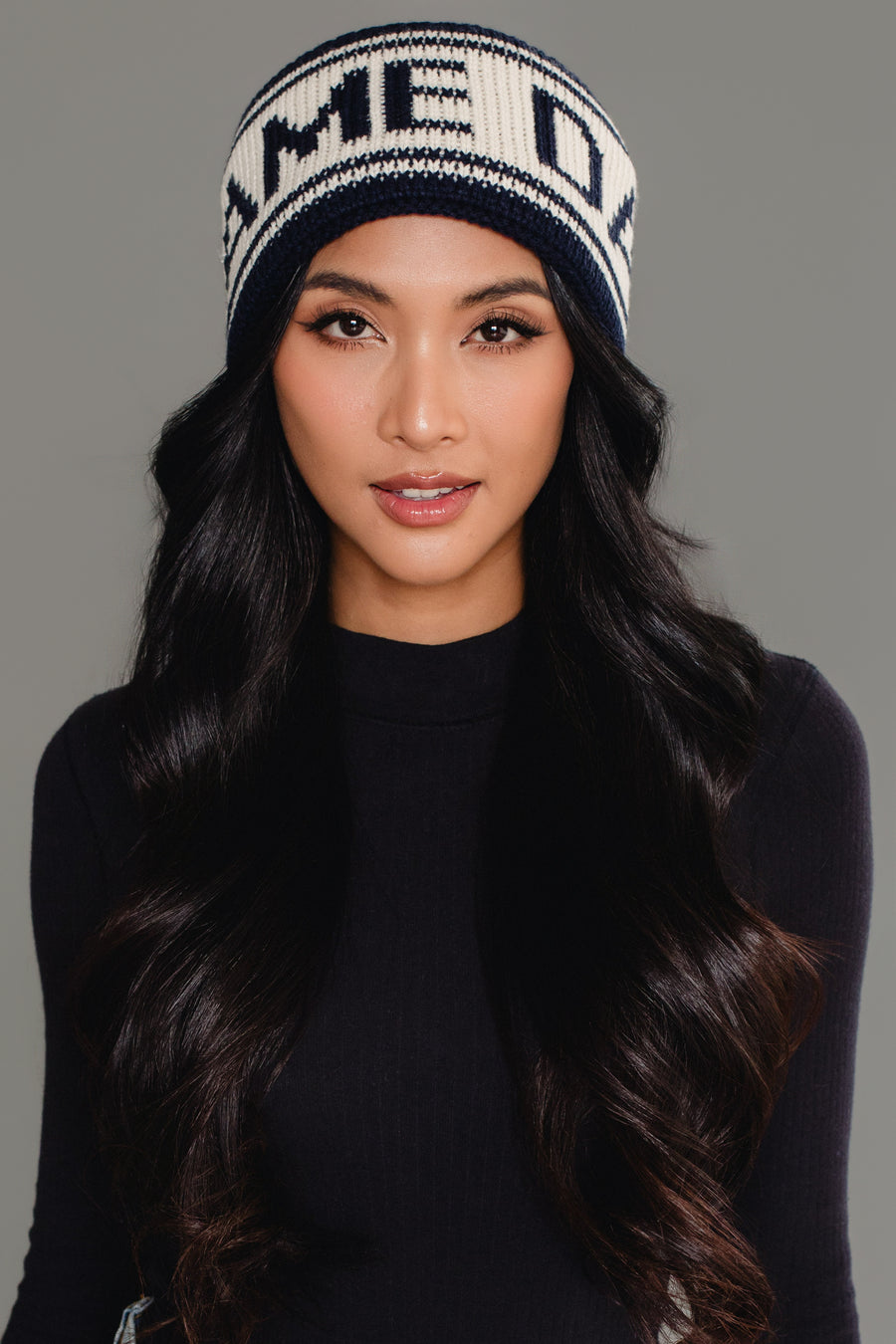 A woman with long wavy black hair, dressed in a black top, wears the Panache Apparel Collection’s Dark Navy & Beige Game Day Knit Headband. The background is plain gray.
