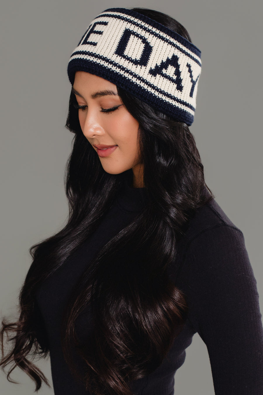 A woman with long dark hair, wearing a black top and the Panache Apparel Collection’s Dark Navy & Beige Game Day Knit Headband featuring bold lettering, looks down with a slight smile against a plain gray background.