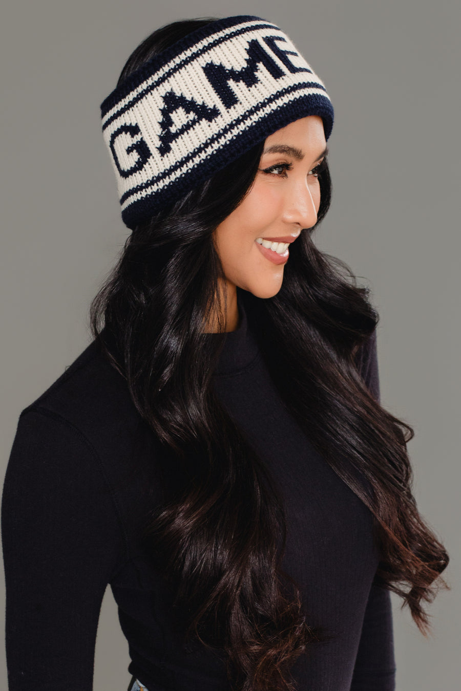 A woman with long dark hair is wearing a black long-sleeve top and the Panache Apparel Collection Dark Navy & Beige Game Day Knit Headband, which features the word "GAME" in bold black letters. She is smiling and looking to her left against a plain gray background.