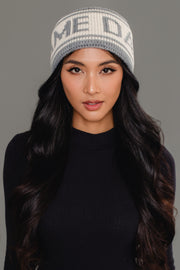 A woman with long dark hair wears the Panache Apparel Collection Gray & Beige Game Day Knit Headband, featuring partially visible text, along with a black long-sleeve top. She stands against a plain gray background.
