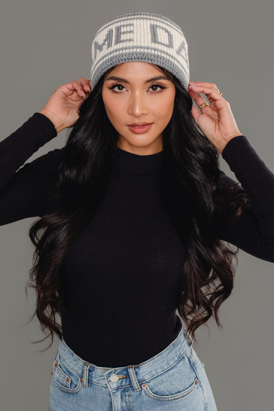 A woman with long black hair wears a black long-sleeve shirt, light blue jeans, and the Panache Apparel Collection Gray & Beige Game Day Knit Headband, posing with both hands holding the sides of her headband against a gray background.