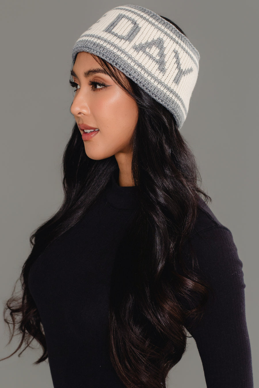 A woman with long dark hair, dressed in a black top and wearing the Panache Apparel Collection Gray & Beige Game Day Knit Headband featuring “DAY” lettering, looks to the side against a plain gray background.