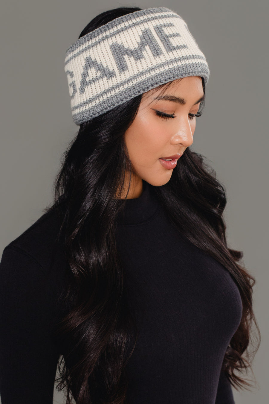 A woman with long black hair wears a black top and the Panache Apparel Collection's Gray & Beige Game Day Knit Headband, featuring the word “GAME,” as she poses against a plain gray background.