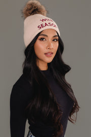 A woman with long dark hair wears the Panache Apparel Collection’s Wine Season Pom Hat, a cream knit hat with a faux fur pom-pom and “WINE SEASON” in red, paired with a black ribbed shirt, posing against a gray background.