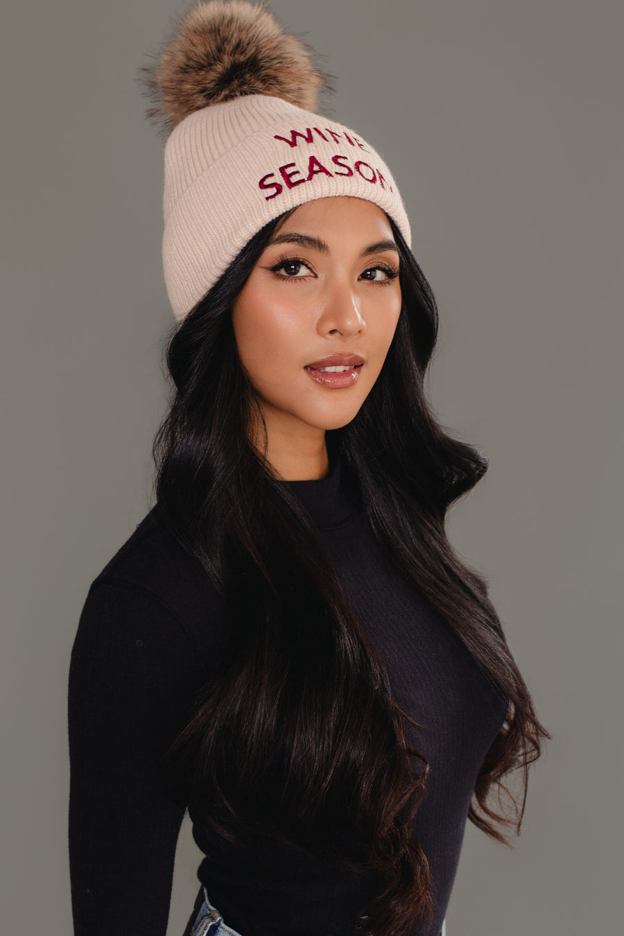 A woman with long dark hair wears the Panache Apparel Collection’s Wine Season Pom Hat, a cream knit hat with a faux fur pom-pom and “WINE SEASON” in red, paired with a black ribbed shirt, posing against a gray background.