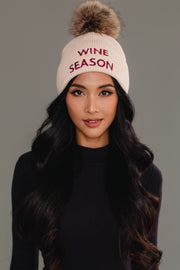 A woman with long black hair is wearing the Panache Apparel Collection’s Wine Season Pom Hat, a beige knit hat featuring a furry pom-pom and “WINE SEASON” embroidered in red, along with a black long-sleeve top, standing against a plain gray background.