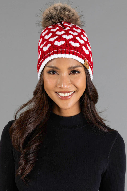 A woman smiles while wearing the Panache Apparel Collection Red & White Heart Pattern Pom Hat, featuring white heart designs and a brown faux fur pom-pom, paired with a black long-sleeve top against a plain gray background.