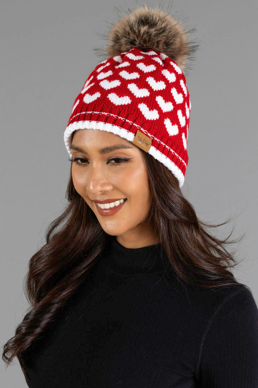 A woman wearing the Panache Apparel Collection Red & White Heart Pattern Pom Hat, featuring white heart designs and a faux fur pom-pom, smiles with long, wavy dark hair and a black top against a gray background.