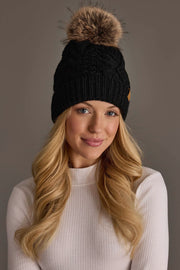 A woman with long blonde hair wears the Panache Apparel Collection Black Cable Knit Pom Hat, featuring a brown faux fur pom and cozy fleece-lined interior, paired with a white ribbed turtleneck as she smiles softly against a plain background.