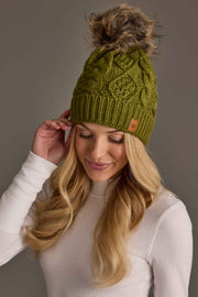 A woman wears an Olive Green Cable Knit Pom Hat in olive green cable knit topped with a brown faux fur pom.