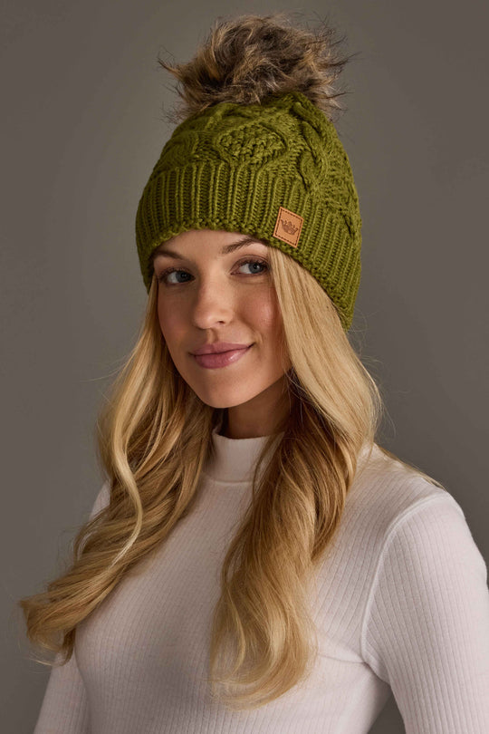 A woman wearing an Olive Green Cable Knit Pom Hat with a faux fur pom-pom.
